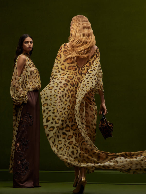 Two models stand against a green backdrop. The model on the right is wearing a jaguar-printed caftan and the model on the right is wearing a jaguar-printed blouse and maroon pants. 