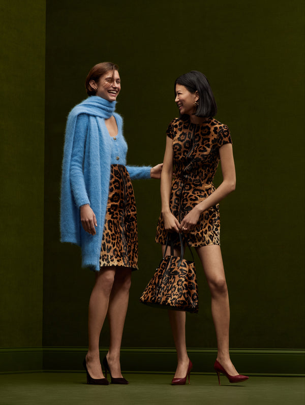 Two models pose together. The model in the left is wearing a blue cardigan and scarf paired with a jaguar pencil skirt, and the model in the right is wearing a short sleeve jaguar mini dress with a matching tote.