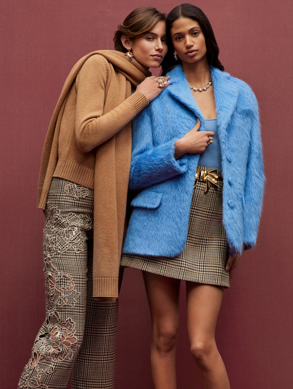 Two models stand next to each other against a pink backdrop. The model on the left is wearing a brown knit sweater & scarf and plaid pants with  peony guipure appliqués and the model on the right is wearing a blue blazer and a plaid skirt with a bow belt. 