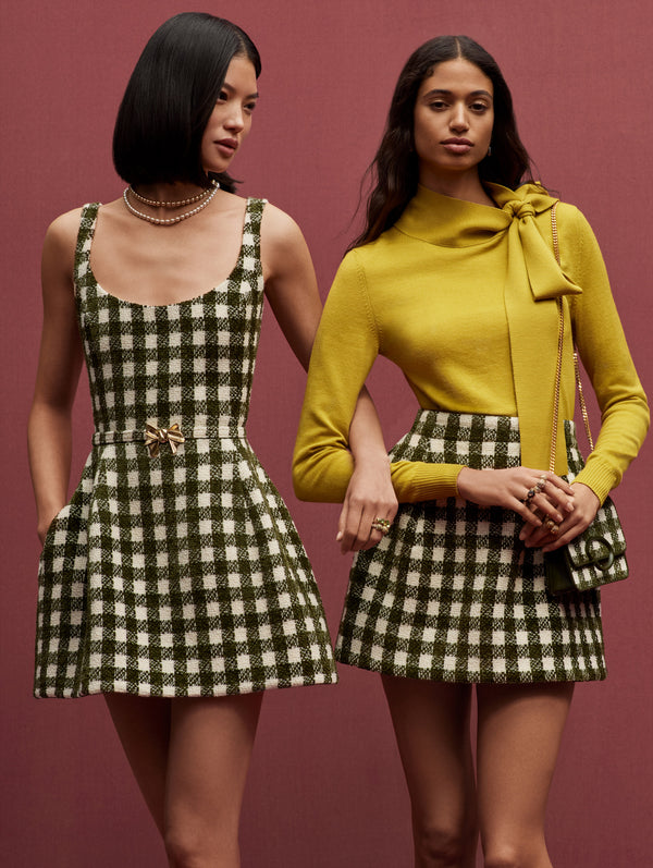 Two models pose in similar outfits against a pink backdrop. The model on the left is in a green and white checkered tweed dress and the model on the right is in a ginger blouse and green and white checkered tweed skirt. 