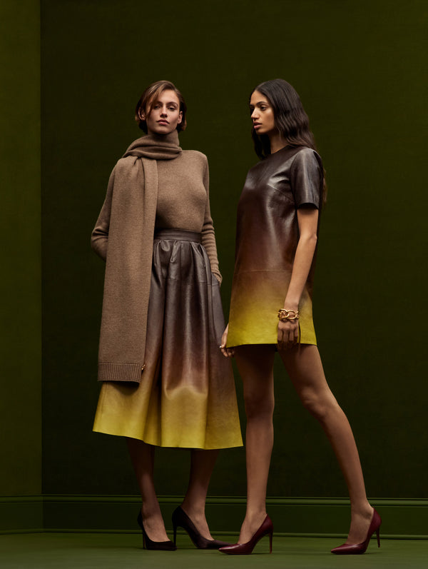 Two models are standing next to each other against a dark green backdrop. The model on the right is in an ombre mini dress and the model on the left is in a chocolate brown ombre skirt with a matching brown sweater and skirt. 