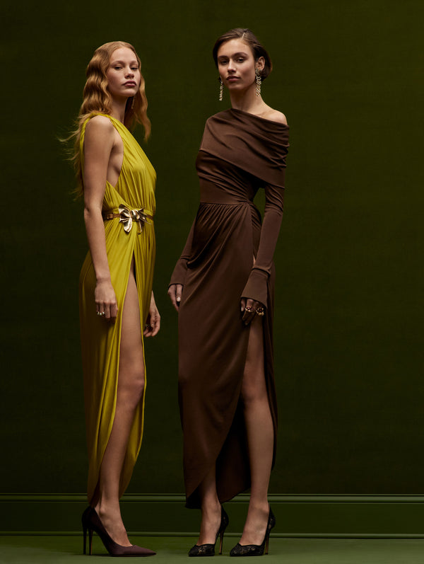 Two models are posing together against a dark green background. The model on the left is wearing a ginger dress and the model on the right is wearing a chocolate off the shoulder jersey dress from Oscar de la Renta's Fall 2024 Collection.