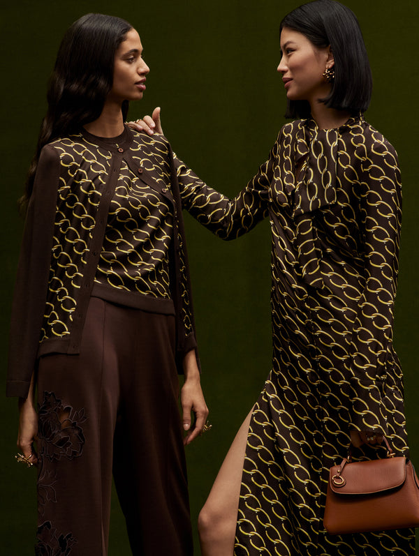 Two models pose together. The model in the left is wearing brown trousers with cutout details in the side paired with an Oscar chain-link tank and matching cardigan. The model in the rights wears a long sleeve dress with the same print while holding the mini Poppy handbag in brown.