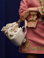 Close up of model in pink mini skirt and jacket with floral embroidered details, while holding the O handle bag in white with three-dimensional roses.