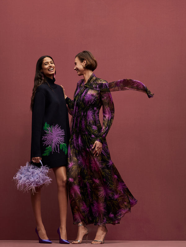 Two models pose together. The model in the left is wearing a navy turtleneck mini dress with a purple dandelion in the side, paired with a purple feathers bag. The model in the right is wearing a long sleeve chiffon dress with purple dandelions.