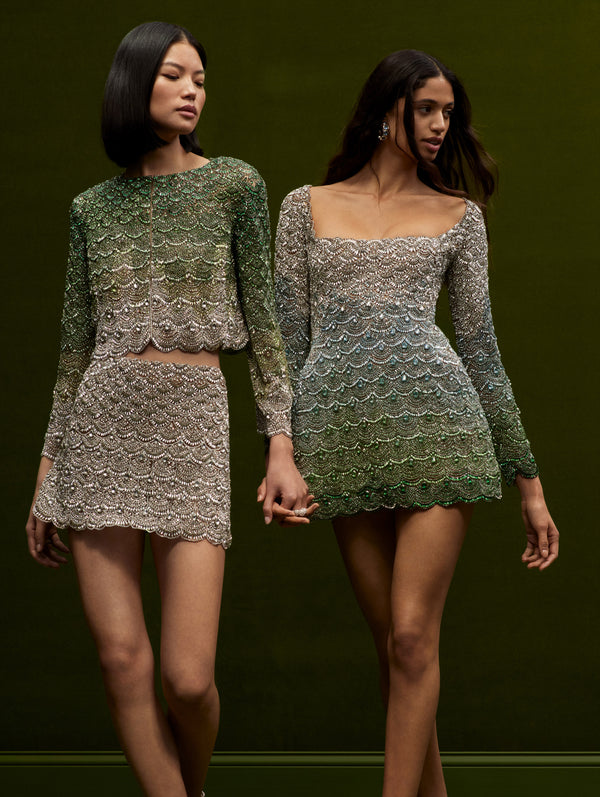 Two models wearing embellished embroidered ombre pieces in tones of green, blue and white. The model in the left wears a mini skirt with the matching long sleeve top, and the model in the right wears a mini long sleeve cocktail dress.