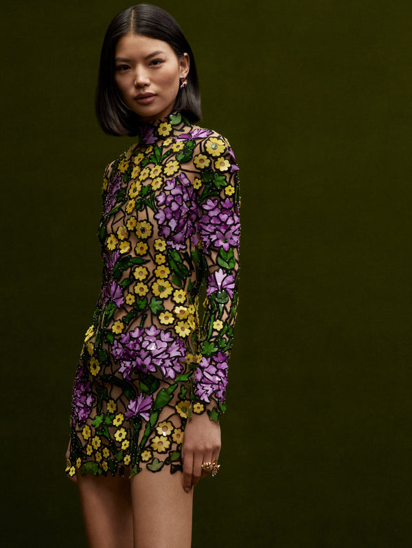 The model wears a long sleeve mock neck mini dress with purple and yellow flowers in mosaic effect.