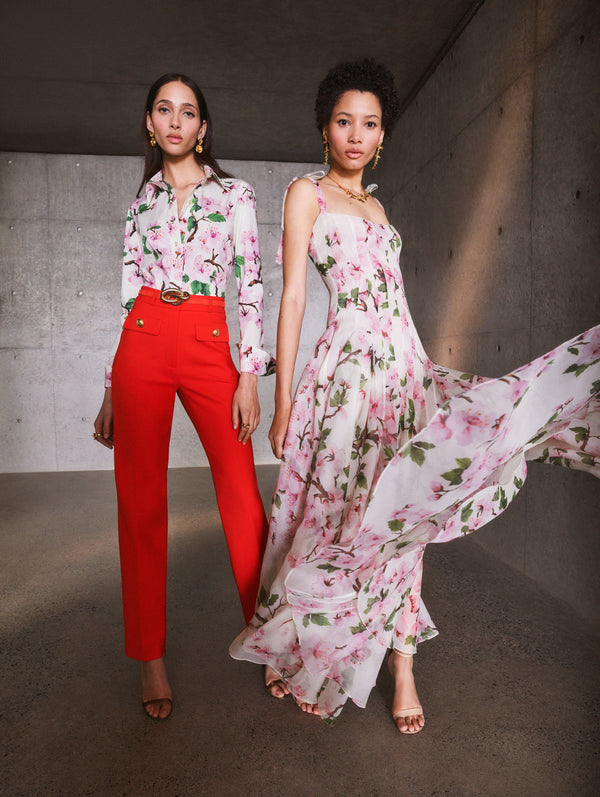 Two models pose in Pre-Fall 2025 looks from Oscar de la Renta’s latest collection. One model wears a floral cotton poplin button down blouse with red pants, and the other wears a floral gown. 