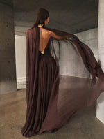 A model poses indoors in a brown gown from Oscar de la Renta’s Pre-Fall 2025 collection. Her back is exposed to the camera detailing butterfly shaped gold hardware. 