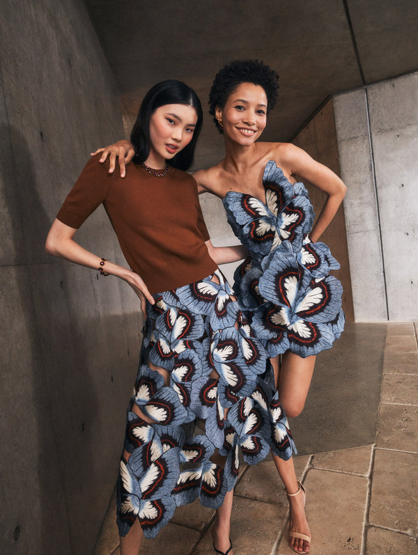 Two models pose in Pre-Fall 2025 looks from Oscar de la Renta’s latest collection. One model wears a brown pullover with a blue and brown butterfly embroidered skirt, and the other model wears a blue and brown butterfly embroidered cocktail dress. 