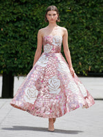 A model is wearing a strapless, tea-length dress with a mosaic-inspired pattern featuring large white roses set against a background of pink and metallic tones. 