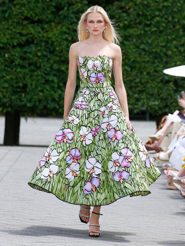 A model is wearing a strapless, A-line dress with a vibrant floral pattern resembling stained glass, featuring large white and pink flowers with green stems. 