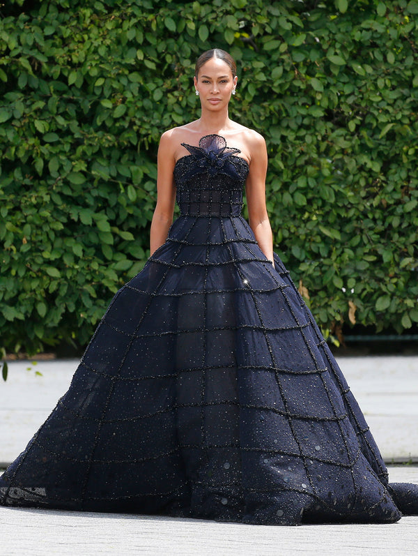 A model is wearing a strapless navy-blue ball gown adorned with subtle sparkles and horizontal seam details, giving it a textured grid pattern. The gown features a large, delicate floral embellishment at the neckline.