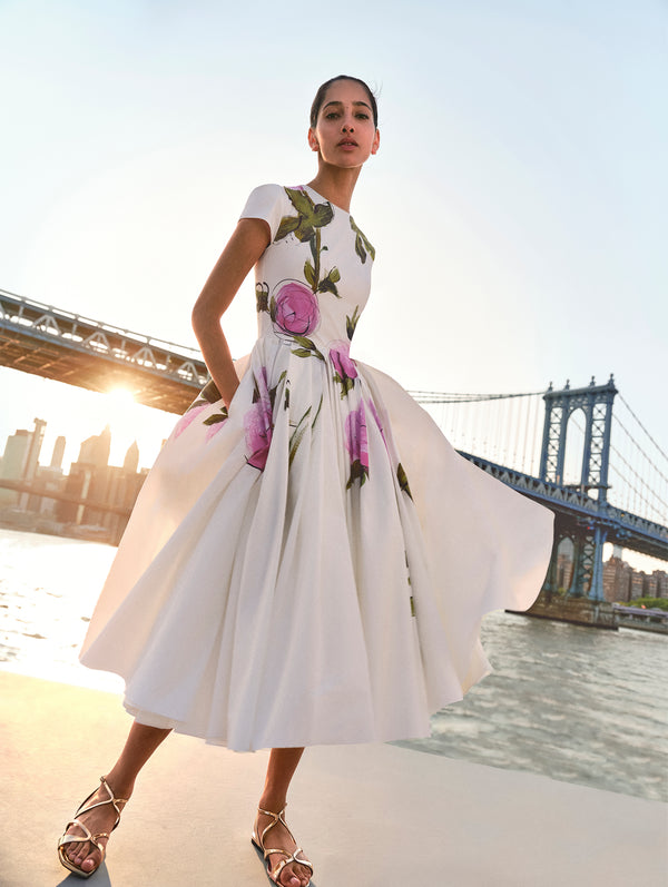 The model poses in a white dress adorned with the pink abstract rose print hand painted by Co-Creative Director Fernando Garcia from Oscar de la Renta's Spring 2025 collection against the New York City skyline.  