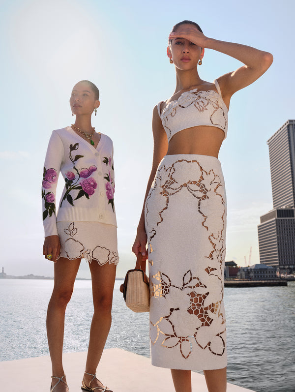 Two models pose next to each other. The model on the left is wearing a long sleeve white cardigan with painted pink roses and a white mini skirt. The model on the right is wearing a matching white top and midi skirt with floral cutouts. 