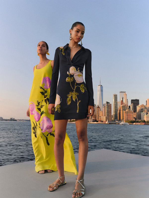 Two models stand on a boat against the New York City backdrop. The model on the right is wearing a short navy floral dress and the model on the left is wearing a long yellow floral dress from Oscar de la Renta's Spring 2025 collection. 