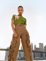 The model is wearing brown high waisted pants with rose cutouts and a green high neck sweater with a pink rose. 