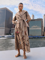 The model poses in a tan trench coat with floral cutout details on the sleeves and at the hem from Oscar de la Renta's Spring 2025 collection against the New York City skyline. 