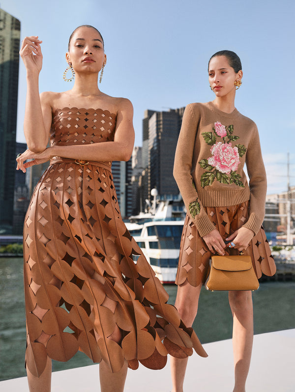 Two models pose neck to each other. The model on the left is wearing a brown leather strapless dress with geometric cutouts. The model on the right is wearing a brown knit pullover sweater with a pink English rose and a brown leather skirt with geometric cutouts. 