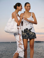 Two models pose next to each other. The model on the left is wearing a white crochet dress with white knit flowers and carrying a matching bag. The model on the right is wearing a white crochet top with a blue and white floral denim mini skirt. 