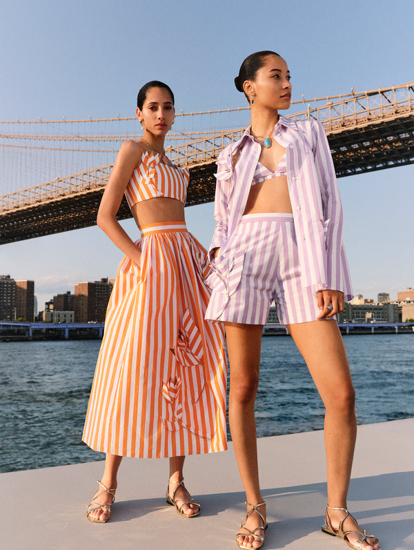 Two models pose next to each other. The model on the left is wearing a matching orange and white striped crop top and skirt. The model on the right is wearing a matching lavender and orange striped button down and shorts set. 