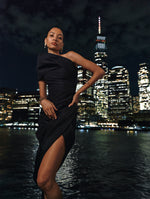 The model is wearing a black dress with draped off-shoulder detailing and an asymmetric hem against the NYC skyline at night. 