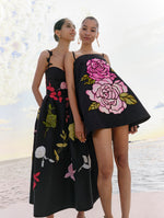 Two models pose standing next to each other. The model on the left is wearing a black midi dress with multicolored flowers. The model on the right is wearing a black mini dress with pink mosaic roses. 