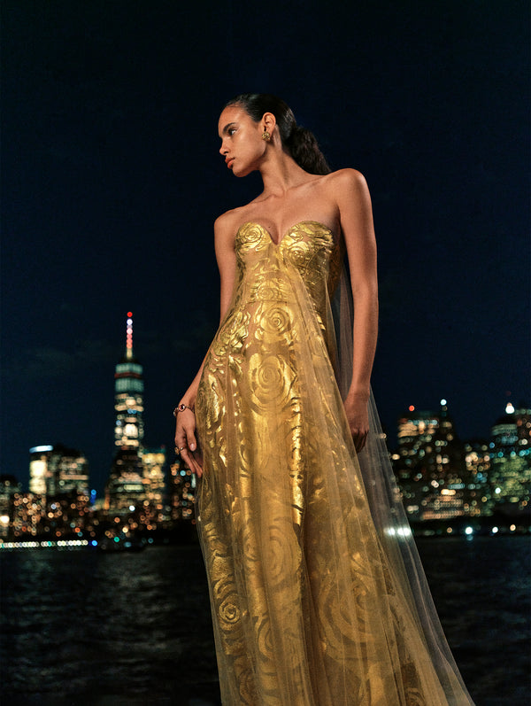 The model poses against the night skyline in a gold gown with rose details from the Oscar de la Renta Spring 2025 collection. 