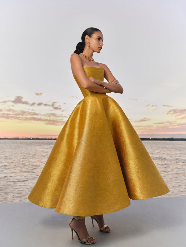 The model is wearing a silk satin yellow gold midi dress from Oscar de la Renta's Spring 2025 Collection against the sunset. 