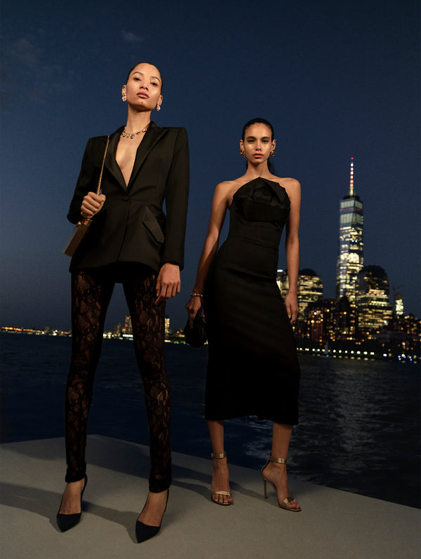 Two models pose against the New York City skyline at night. The model on the left is wearing lace pants and a black tuxedo jacket and the model on the left is wearing a black midi cocktail dress with a rose at the bust from Oscar de la Renta's Spring 2025 collection. 