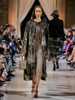 Holding a gold sequin-embroidered cube-like handbag, the model walks the runway in a brown open-front long-sleeve fur coat, layered over a metallic semi-sheer mesh dress with threadwork embroidered leaves. Behind her, the blurred background shows the audience and several models walking the runway in various directions.