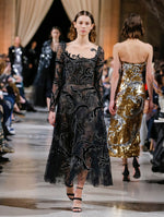 The model walks the runway in a black semi-sheer illusion-neck long-sleeve cocktail dress with swirling black and silver embroidery. Behind her, the blurred background shows the audience and several models walking the runway.